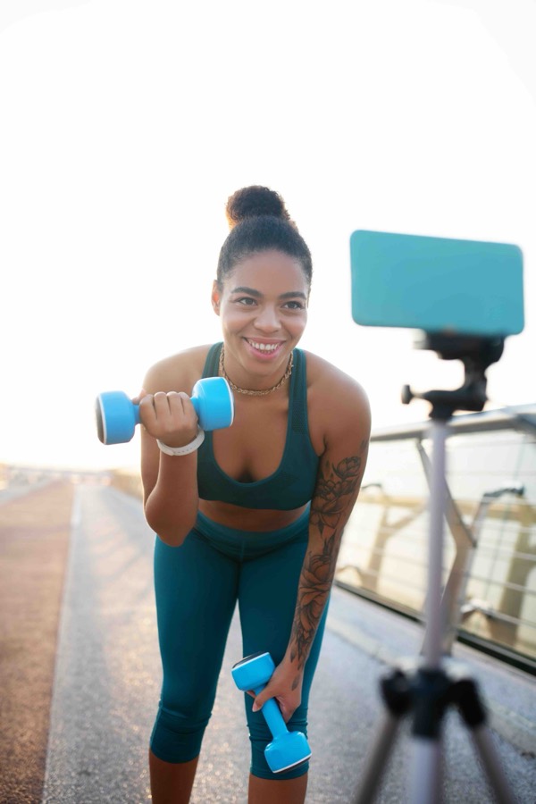 A creator in teal activewear curling a dumbbell while filming on a phone tripod at sunrise