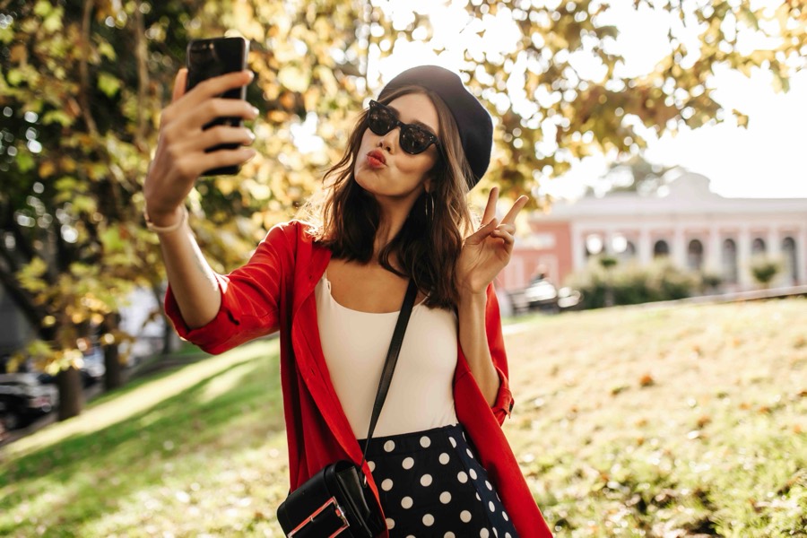 A creator in a red jacket and black beret taking a selfie on her phone in a sunlit park