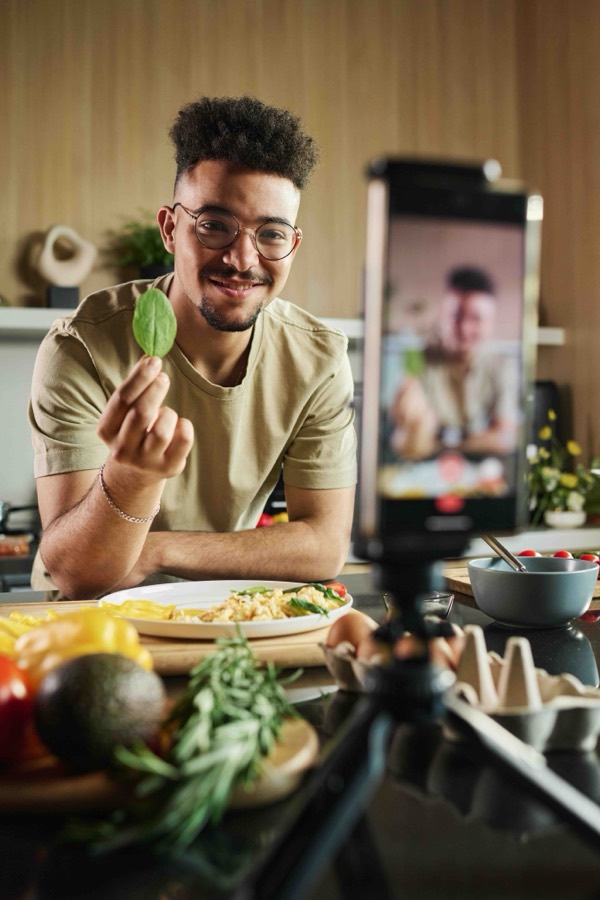 A food creator holding up a spinach leaf while filming a plated dish on a phone tripod in a kitchen