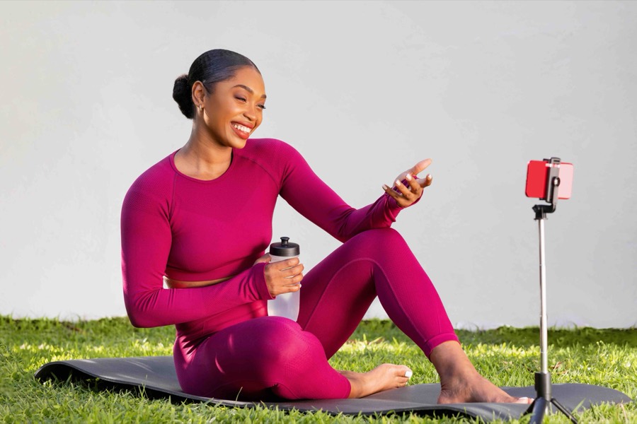 A creator in magenta activewear filming herself on a phone clipped to a selfie stick while sitting on a yoga mat on grass