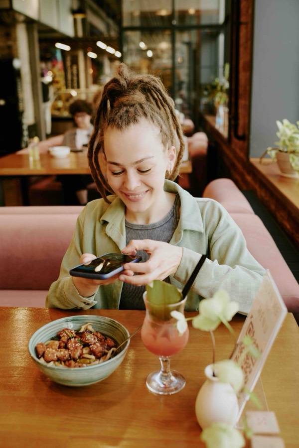A diner photographing a noodle bowl and a cocktail on her phone at a restaurant table